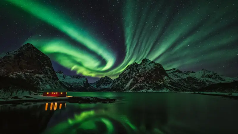 Green northern lights over a red cabin beside a Norwegian fjord in winter