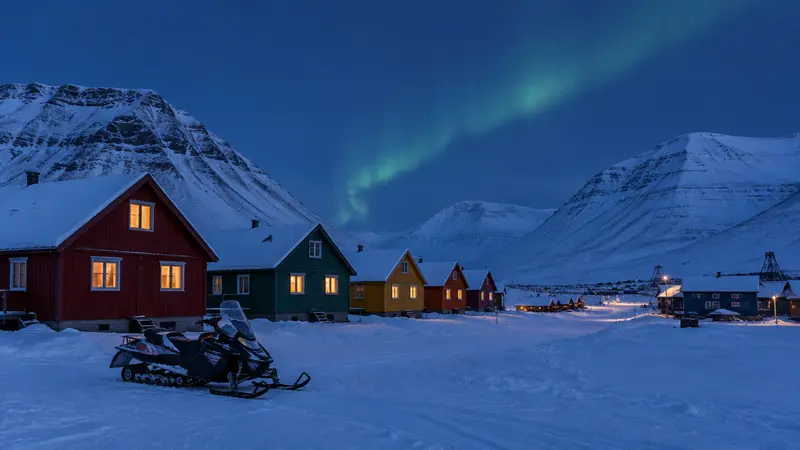 Northern lights over colourful wooden houses and a parked snowmobile in a snow-covered Arctic valley during the polar night
