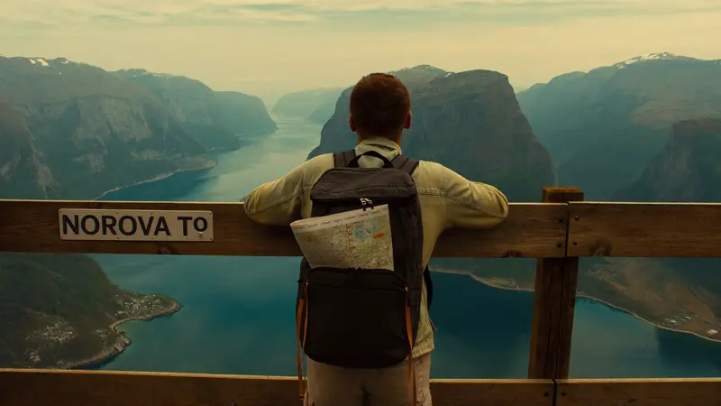 Traveler with a backpack at a wooden viewpoint rail overlooking layered fjord ridges